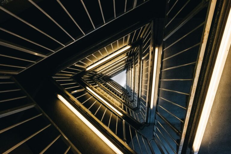 A dynamic, high-angle downward perspective of a triangular staircase. Warm fluorescent lights illuminate each level, creating an infinite, converging vortex. This image illustrates the triangle's core meanings of direction, infinite transformation, and structural rigidity as discussed in the article.