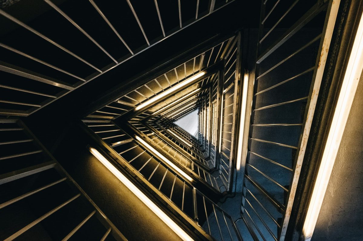A dynamic, high-angle downward perspective of a triangular staircase. Warm fluorescent lights illuminate each level, creating an infinite, converging vortex. This image illustrates the triangle's core meanings of direction, infinite transformation, and structural rigidity as discussed in the article.