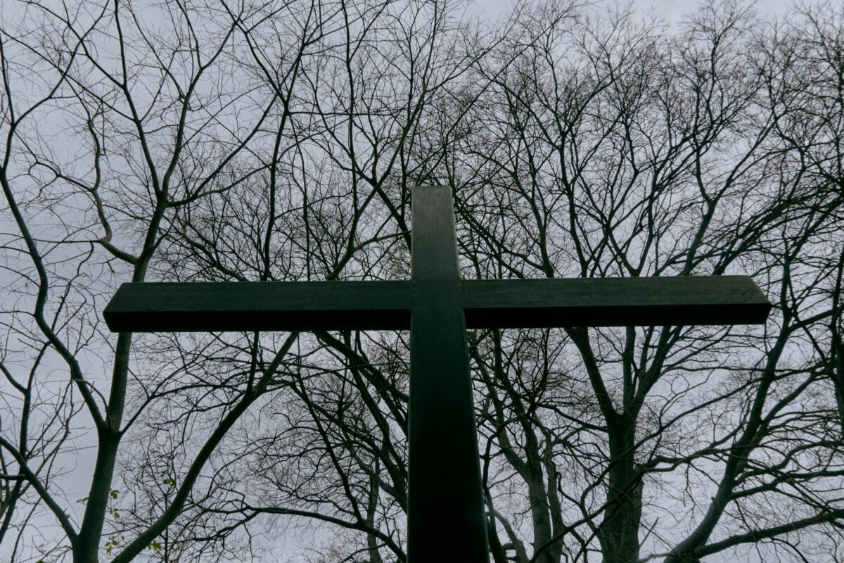 A dramatic, low-angle photograph taken from the base of a large, dark-silhouetted cross, looking up against a dense network of bare, tangled tree branches under an overcast grey sky. The cross is massive, dominating the frame.