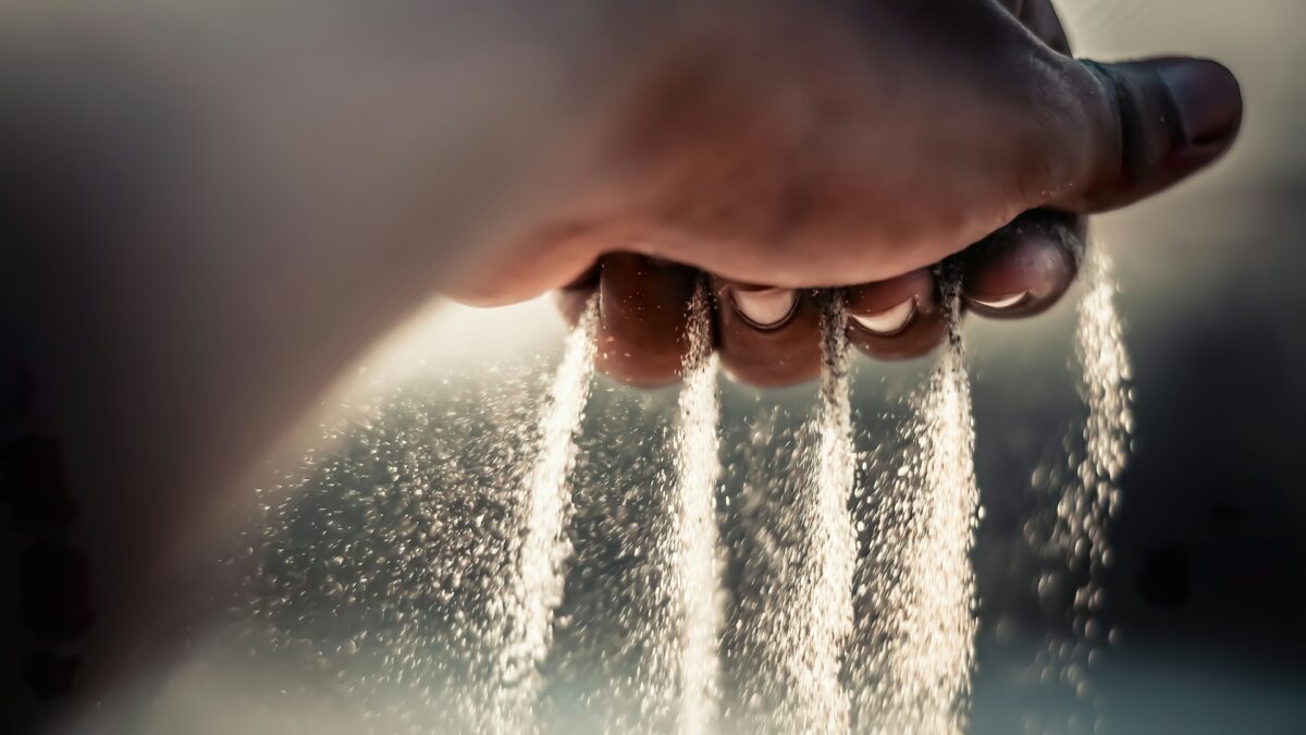 Close-up of white salt crystals falling through a person's fingers with warm backlighting and a bokeh background.