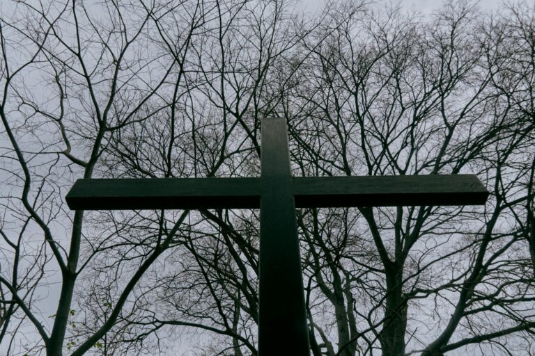 A dramatic, low-angle photograph taken from the base of a large, dark-silhouetted cross, looking up against a dense network of bare, tangled tree branches under an overcast grey sky. The cross is massive, dominating the frame.